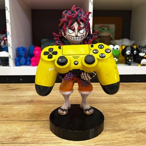 May include: A yellow PlayStation 4 controller is held by a figurine of a cartoon character with red hair and a menacing grin. The figurine is standing on a black circular base.