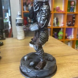 May include: A detailed Predator figurine, predominantly silver and brown. The figure is posed on a grey base decorated with skulls. The Predator is shown in a dynamic pose, with detailed armour and weaponry. A collectible item, suitable for display.