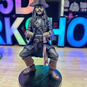 May include: A detailed figurine of Captain Jack Sparrow from the Pirates of the Caribbean film series. The figure is in a dynamic pose, wearing a brown tricorn hat, a long coat, and boots. The base is black and round.