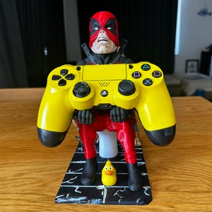 May include: A yellow PlayStation 4 controller is held by a 3D printed figurine of Deadpool sitting on a toilet. The figurine is on a black and white base with a yellow rubber ducky in front of it.