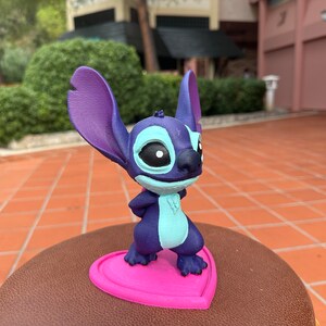 May include: A blue and purple Stitch figurine standing on a pink heart-shaped base. The figurine is a popular Disney character.