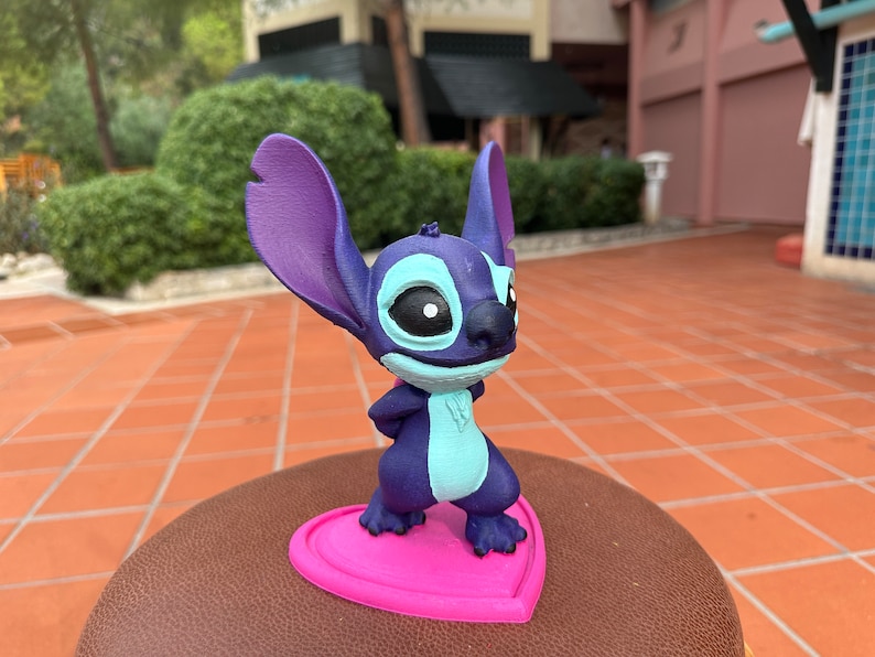May include: A blue and purple Stitch figurine standing on a pink heart-shaped base. The figurine is a popular Disney character.
