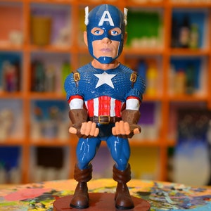 May include: A blue and red Captain America figurine with a white star on his chest. He is wearing a blue helmet with a white 'A' on it. The figurine is standing on a brown base.