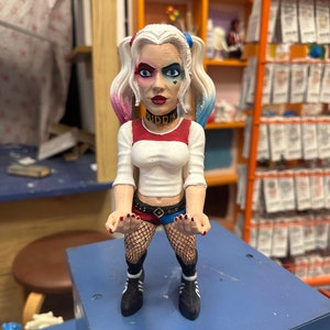 May include: A hand-painted figurine of Harley Quinn, a DC Comics character. The figurine is wearing a red and black costume with a white shirt and black fishnet stockings. The figurine has a black choker with the word "PUDDIN" written on it. The figurine is standing on a blue surface.