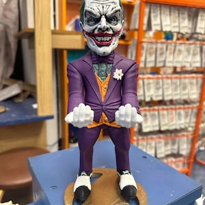 May include: A detailed figurine of the Joker, wearing a purple suit with an orange waistcoat and a green shirt. The figure has white gloves, black and white shoes, and a gold-coloured base. The Joker's face is painted with white, red, and green details.