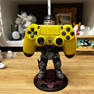 May include: A yellow PlayStation 4 controller is held by a black and grey action figure. The figure is standing on a black base with a red and black design.