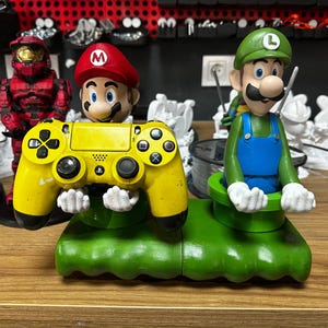May include: A yellow and black video game controller is held by a green and red Mario character figurine. The controller is mounted on a green and brown base. A green and blue Luigi character figurine is holding a green pipe on the right side of the base.