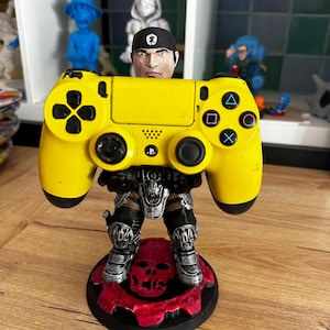 May include: A yellow PlayStation 4 controller is held by a 3D printed figurine of a soldier wearing a black bandana and armour. The figurine is standing on a circular base with a red skull and gears design.