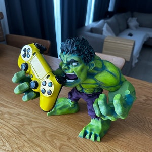 Könnte beinhalten: Eine grüne Hulk-Figur, die einen gelben Videospiel-Controller hält. Die Figur ist aus Kunststoff und hat eine detaillierte Bemalung. Der Controller ist ein PlayStation 4-Controller.