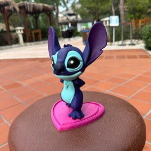 May include: A blue and purple 3D printed figurine of Stitch from Lilo & Stitch standing on a pink heart-shaped base.