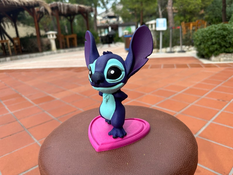 May include: A blue and purple 3D printed figurine of Stitch from Lilo & Stitch standing on a pink heart-shaped base.