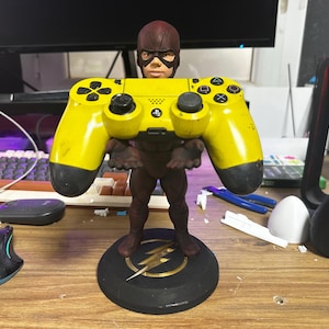 Puede incluir: Una figura de The Flash sosteniendo un mando de PlayStation amarillo. La figura es marrón y roja, de pie sobre una base negra con un emblema de rayo dorado. El mando es amarillo con botones negros.