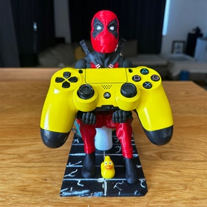 May include: A yellow PlayStation 4 controller is held by a 3D printed figurine of Deadpool sitting on a toilet. A yellow rubber ducky sits in front of the figurine.