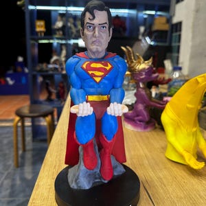 May include: A blue and red Superman figurine with a black base. The figurine is sitting down with its arms crossed and has a serious expression on its face. The base of the figurine has the word "SUPERMAN" written on it.