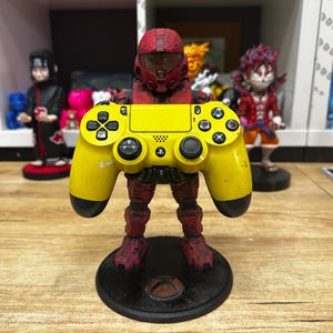 以下が含まれることがあります： 黄色のプレイステーション4コントローラーを、赤と黒のフィギュアが持っています。フィギュアは黒い円形の台座の上に立っています。