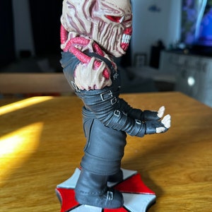 Pode incluir: Uma figura detalhada de um personagem monstruoso da s&eacute;rie de videogames Resident Evil. A figura est&aacute; em p&eacute; sobre uma base com o ic&ocirc;nico logotipo da Umbrella Corporation. O personagem tem uma cabe&ccedil;a grotesca e carnuda com olhos vermelhos e dentes afiados. A figura est&aacute; vestindo um traje preto com tiras e fivelas.