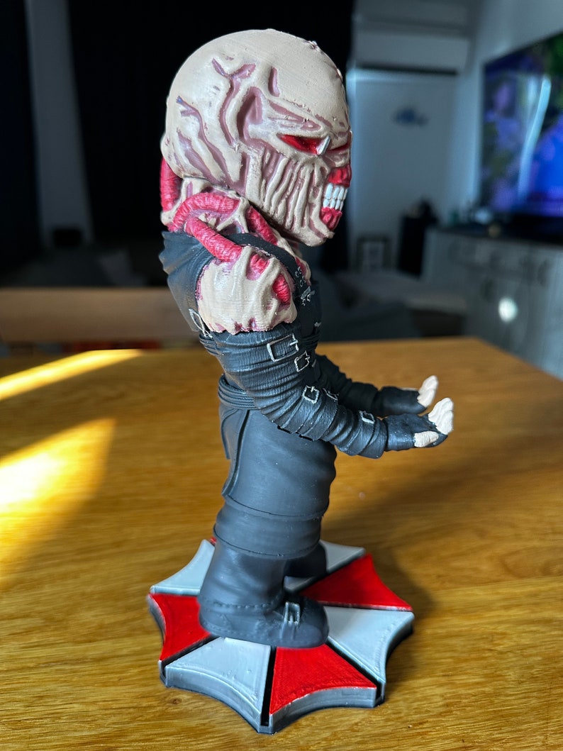 Pode incluir: Uma figura detalhada de um personagem monstruoso da s&eacute;rie de videogames Resident Evil. A figura est&aacute; em p&eacute; sobre uma base com o ic&ocirc;nico logotipo da Umbrella Corporation. O personagem tem uma cabe&ccedil;a grotesca e carnuda com olhos vermelhos e dentes afiados. A figura est&aacute; vestindo um traje preto com tiras e fivelas.