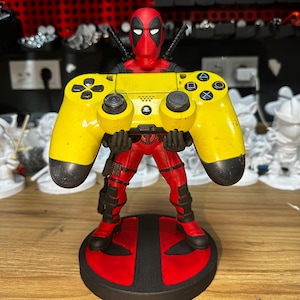 May include: A yellow and black video game controller held by a red and black Deadpool figurine. The figurine is standing on a black and red base with the Deadpool logo.
