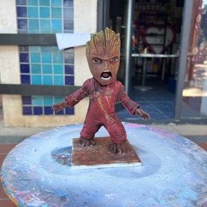 May include: A small, brown, tree-like figure with a red jumpsuit and a zipper. The figure has a green, leafy head and is standing on a brown base. The figure is painted with a distressed look, giving it a weathered appearance.