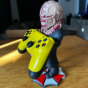 Pode incluir: Uma figura de um personagem zumbi com uma base de guarda-chuva vermelho e branco segurando um controle de PlayStation amarelo. O personagem est&aacute; vestindo uma jaqueta preta e tem um rosto grotesco e detalhado.