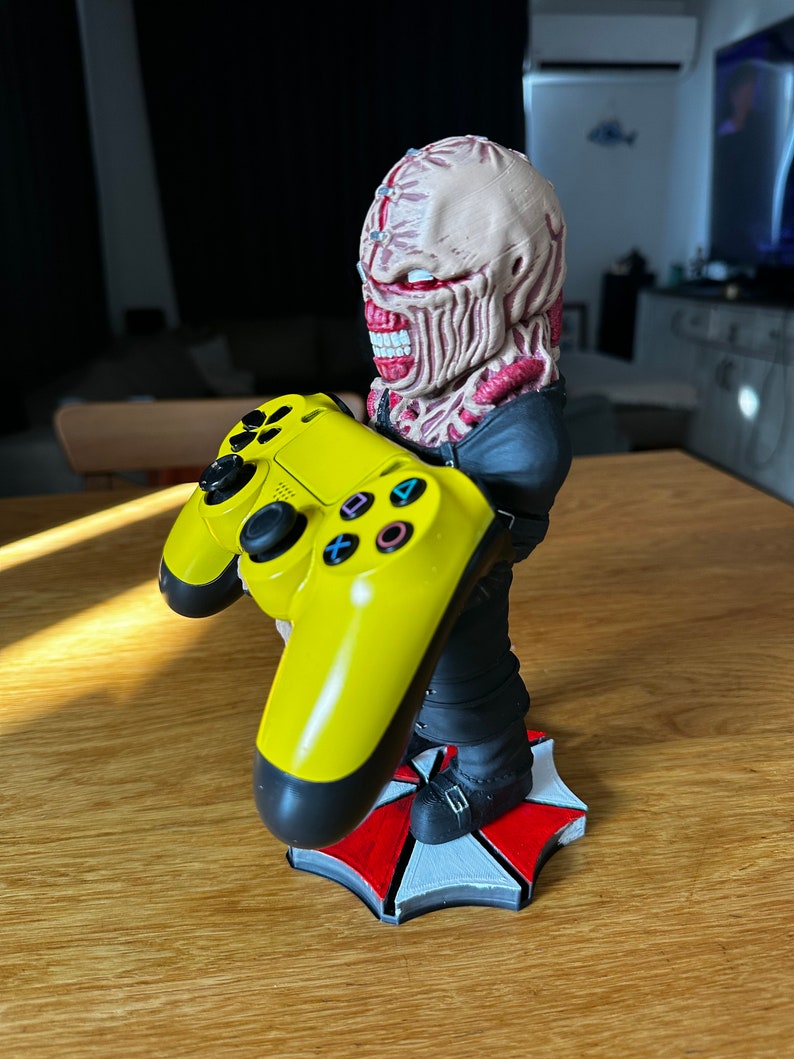 Pode incluir: Uma figura de um personagem zumbi com uma base de guarda-chuva vermelho e branco segurando um controle de PlayStation amarelo. O personagem est&aacute; vestindo uma jaqueta preta e tem um rosto grotesco e detalhado.