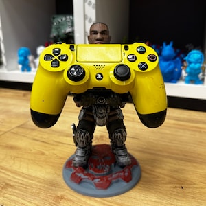 May include: A yellow PlayStation 4 controller held by a detailed figurine. The figurine has a gray and silver armored design and a red skull base. The controller has black buttons and directional pads.