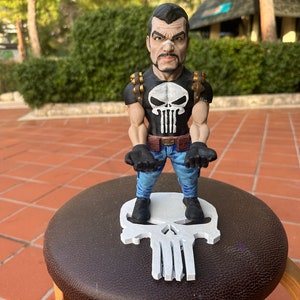 May include: A figurine of a muscular character with a stern expression, wearing a black shirt with a white skull emblem, blue jeans, and black gloves. The figure stands on a white skull-shaped base.