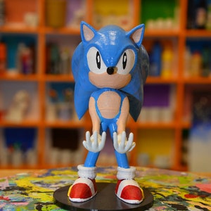 Puede incluir: Figurita de Sonic the Hedgehog impresa en 3D en azul y blanco, de pie sobre una base negra. La figurita está en posición de brazos extendidos y se coloca sobre una superficie pintada de colores.