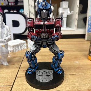 Könnte beinhalten: Detaillierte Figur von Optimus Prime, dem Anführer der Autobots, in einer dynamischen Pose. Die Figur ist überwiegend rot, blau und silberfarben, mit aufwendigen Details. Sie steht auf einer schwarzen Basis mit dem Autobot-Symbol.