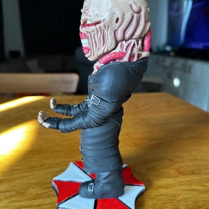 Pode incluir: Uma figura vestida de preto com uma cabe&ccedil;a grotesca, carnuda e de olhos vermelhos est&aacute; apoiada em uma base com o logotipo da Umbrella Corporation, vermelho e branco. A figura tem uma express&atilde;o amea&ccedil;adora e parece ser um personagem de terror.