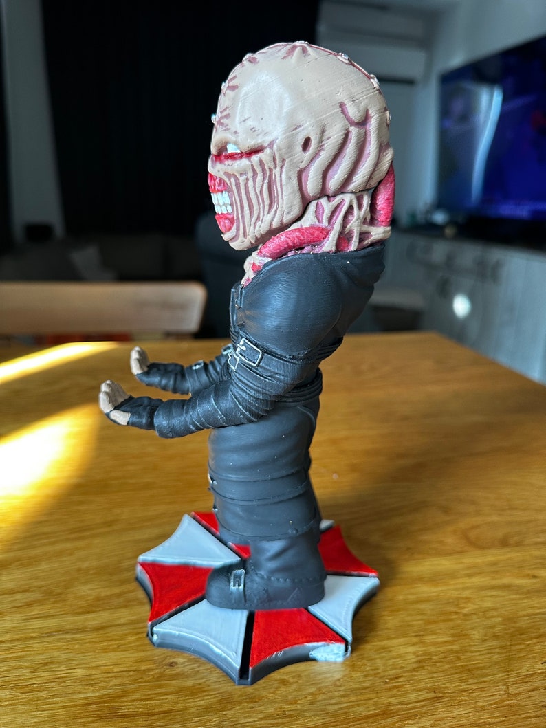 Pode incluir: Uma figura vestida de preto com uma cabe&ccedil;a grotesca, carnuda e de olhos vermelhos est&aacute; apoiada em uma base com o logotipo da Umbrella Corporation, vermelho e branco. A figura tem uma express&atilde;o amea&ccedil;adora e parece ser um personagem de terror.