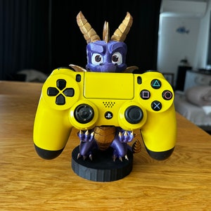 May include: A yellow PlayStation 4 controller is held by a purple and gold dragon figurine. The dragon is holding the controller in its front paws. The controller is on a black base.