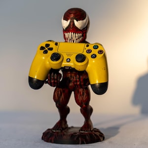 以下が含まれることがあります： 歯を見せて笑っている赤と黒のキャラクターのフィギュアが、黄色のPlayStation 4コントローラーを持っている