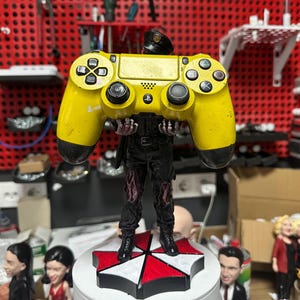 May include: A yellow PlayStation 4 controller held by a figurine. The figurine is dressed in a black uniform with red accents on the legs and a black hat. The base is a red, white, and black geometric design. The background is a workshop setting.