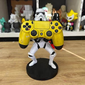 Puede incluir: Una figura de Stormtrooper blanco sobre una base circular negra, sosteniendo un mando de videojuego amarillo brillante con detalles en negro. El mando cuenta con botones direccionales estándar, joysticks y un botón central con el logo de PlayStation.