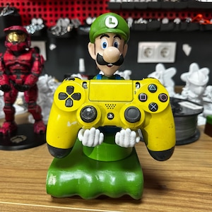 May include: A yellow PlayStation 4 controller is held by a green and white figurine of a video game character. The character is wearing a green hat with an 'L' on it. The controller is sitting on a green base.