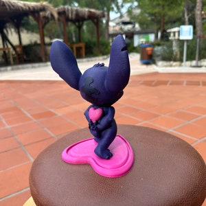 May include: A blue 3D printed figurine of Stitch from Lilo & Stitch holding a pink heart. The figurine is standing on a pink heart-shaped base.