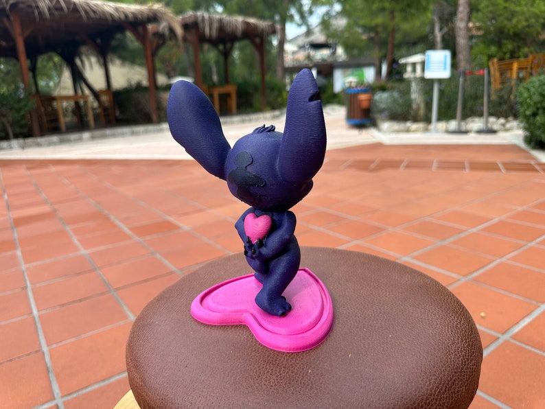May include: A blue 3D printed figurine of Stitch from Lilo & Stitch holding a pink heart. The figurine is standing on a pink heart-shaped base.
