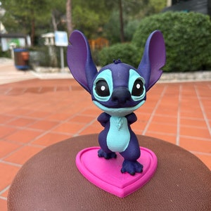 May include: A blue and purple Stitch figurine standing on a pink heart-shaped base. The figurine is made of plastic and has a cartoonish appearance.