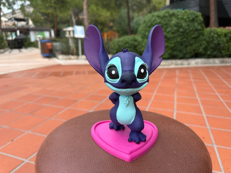 May include: A blue and purple Stitch figurine standing on a pink heart-shaped base. The figurine is made of plastic and has a cartoonish appearance.
