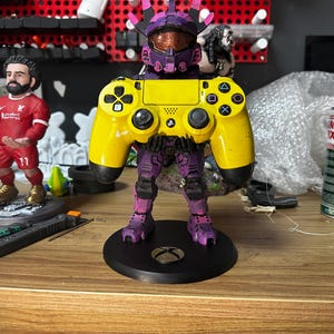 May include: A yellow PlayStation 4 controller is held by a purple and black action figure. The figure is standing on a black circular base.