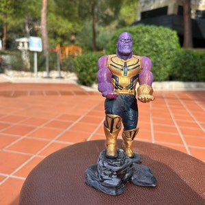 May include: A purple and gold action figure of Thanos, a supervillain from the Marvel Cinematic Universe, standing on a rocky base. Thanos is holding the Infinity Gauntlet in his right hand and a purple gem in his left hand.