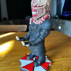 Pode incluir: Uma figura impressa em 3D de um personagem da s&eacute;rie de videogames Resident Evil. O personagem est&aacute; vestindo uma roupa preta com al&ccedil;as e tem um rosto vermelho e branco com uma boca grande. A figura est&aacute; em p&eacute; sobre um logotipo de guarda-chuva vermelho e branco.