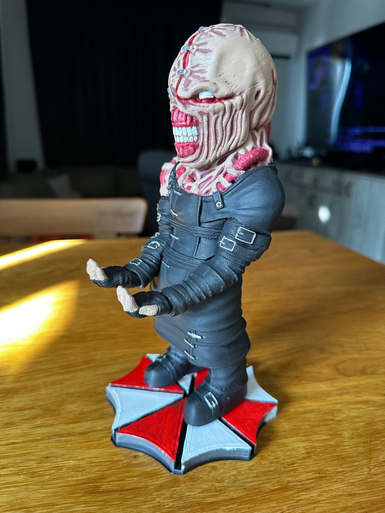Pode incluir: Uma figura impressa em 3D de um personagem da s&eacute;rie de videogames Resident Evil. O personagem est&aacute; vestindo uma roupa preta com al&ccedil;as e tem um rosto vermelho e branco com uma boca grande. A figura est&aacute; em p&eacute; sobre um logotipo de guarda-chuva vermelho e branco.