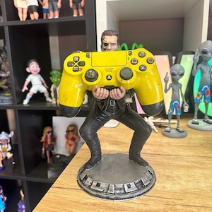Puede incluir: Una figura de un hombre sosteniendo un gran mando de PlayStation amarillo. La figura lleva un atuendo negro y se encuentra sobre una base circular. Otras figuras son visibles en estanterías en el fondo.