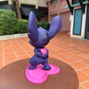 May include: A purple 3D printed figurine of Stitch from Lilo & Stitch holding a pink heart. The figurine is standing on a pink heart-shaped base.