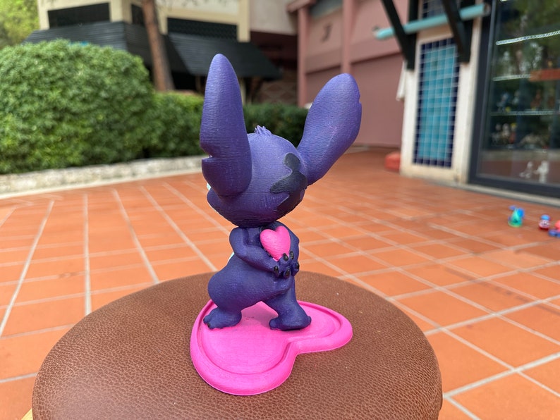 May include: A purple 3D printed figurine of Stitch from Lilo & Stitch holding a pink heart. The figurine is standing on a pink heart-shaped base.