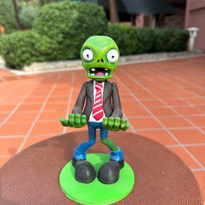 Peut inclure: Une figurine de zombie verte portant un costume noir et une cravate rouge et blanche à rayures. Le zombie a une expression surprise sur son visage et se tient sur une base verte.