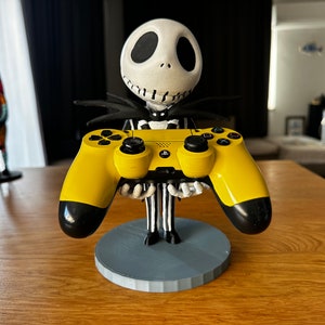 May include: A yellow PlayStation 4 controller is held by a 3D printed figurine of Jack Skellington from the Nightmare Before Christmas. The figurine is standing on a gray circular base.