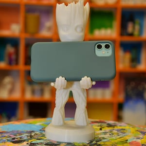May include: A white 3D printed Groot figurine holding a teal phone case with a dual camera lens. The figurine is standing on a colorful paint palette.